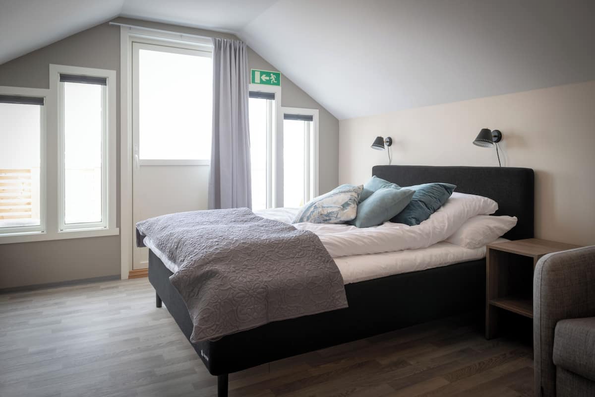 Un dormitorio con cama, dos mesitas de noche, una mesita auxiliar, una ventana con cortinas y una señal de salida verde.