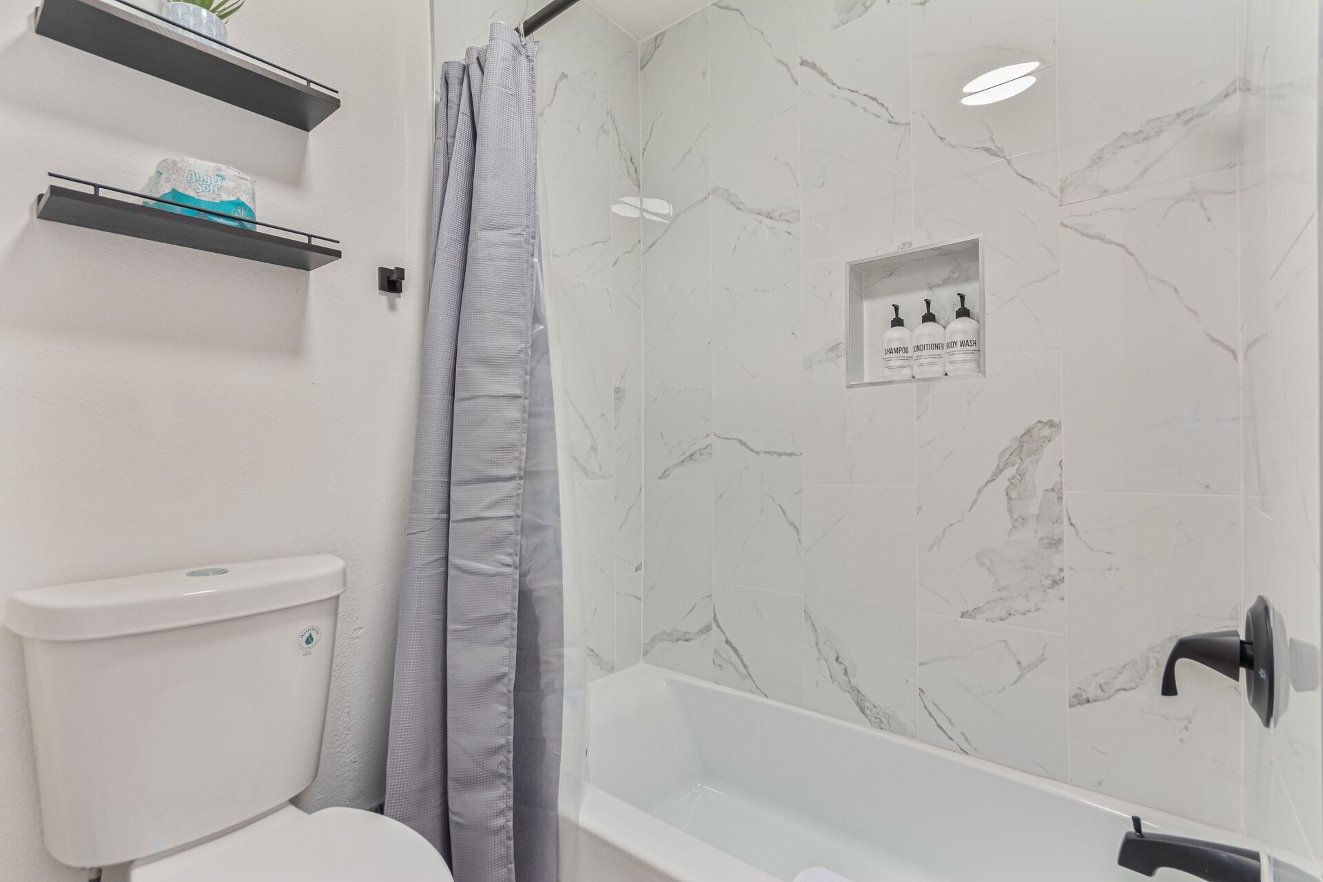 A bathroom with a toilet, a shower with a grey curtain, and a bathtub. There are shelves with toiletries and a sign indicating the bathroom products.