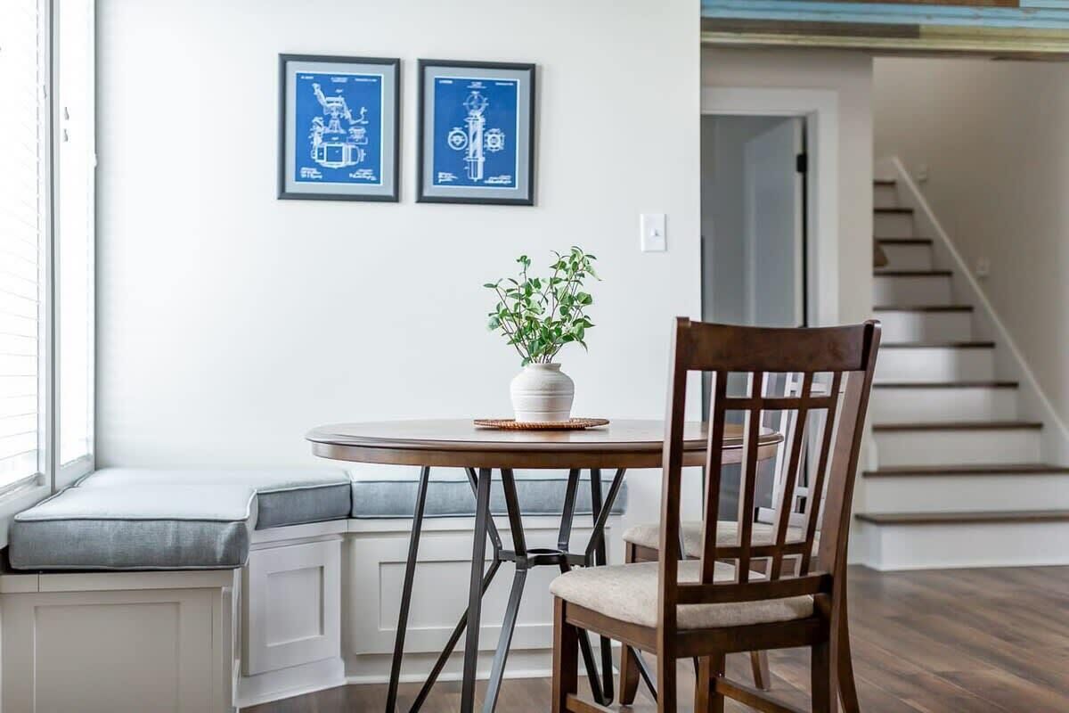 A dining area with a round table, a wooden chair, a bench with a cushioned seat, and two framed blue architectural drawings on the wall.