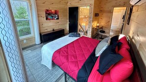 A wooden cabin bedroom with a bed, a ceiling fan, a painting, and a view of the outdoors.