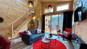 A wooden cabin interior with a sofa, red chairs, a TV, and a staircase.