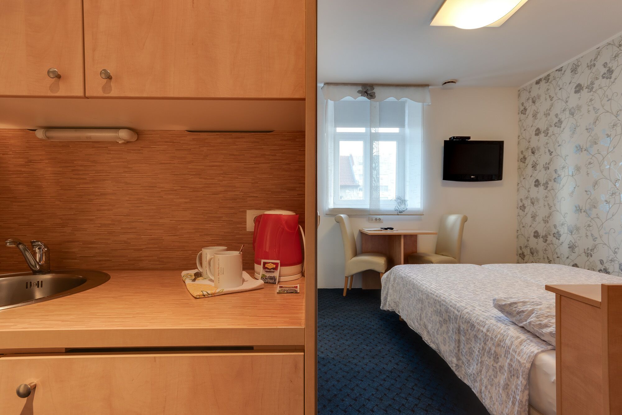 Una habitación de hotel compacta con kitchenette, una cama individual, un escritorio pequeño y un televisor.