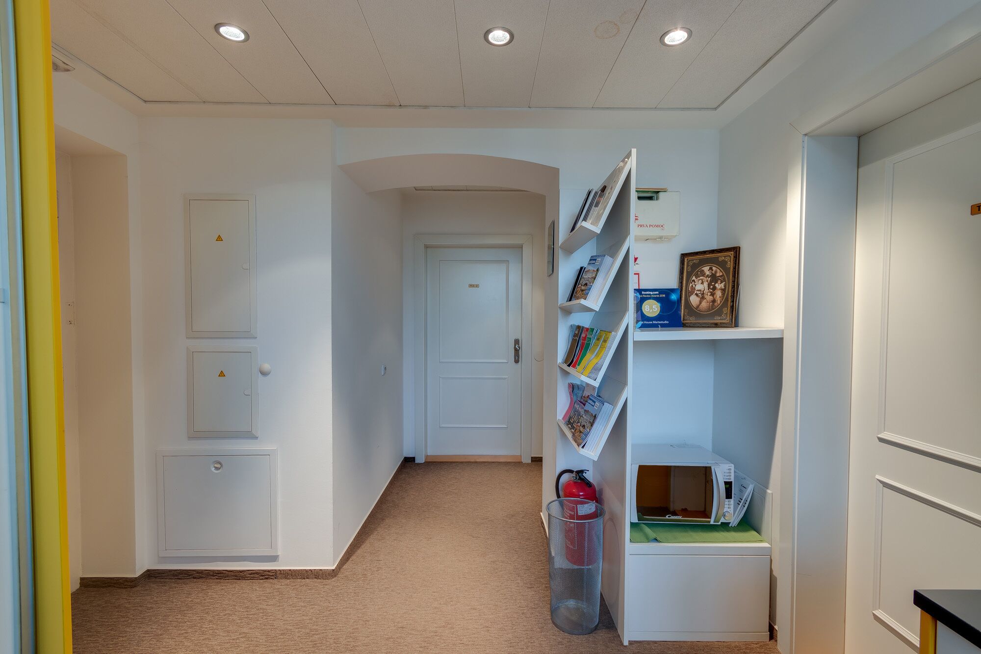A hallway with white walls, a carpeted floor, and a fire extinguisher. There are bookshelves with books and a microwave on a counter.
