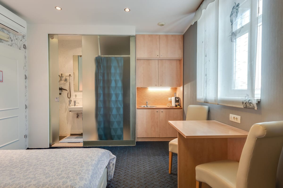 Ein kleines Hotelzimmer mit Bett, einer kompakten Küchenzeile, einer Dusche und einem Fenster mit Vorhängen.