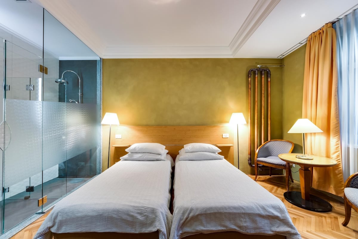 Ein Hotelzimmer mit zwei Betten, einer Duschkabine aus Glas, einem Holz-Kopfteil, einem kleinen runden Tisch und einem Sessel.