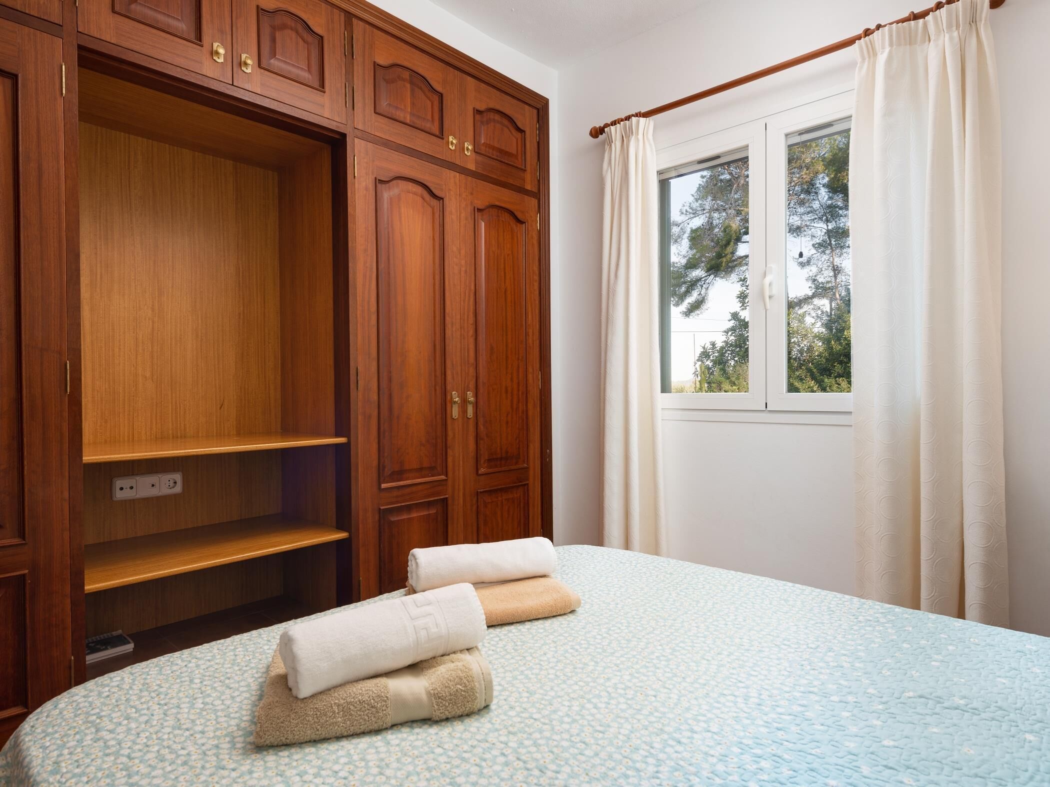 A bedroom with a large wooden wardrobe, a bed with a floral bedspread, and a window with curtains.