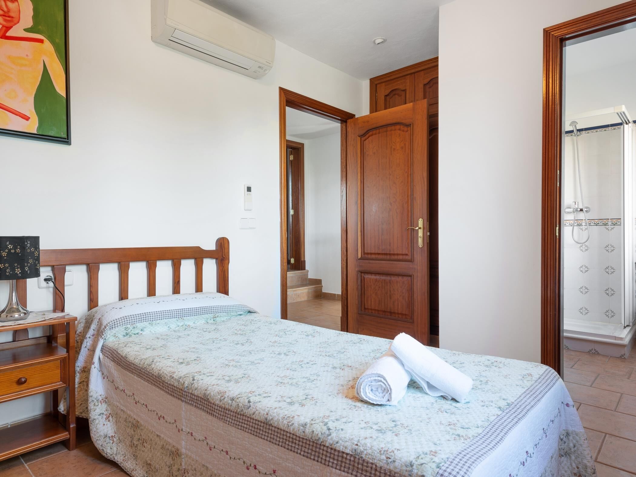 A bedroom with a bed, a nightstand, a wooden door, and a shower area.