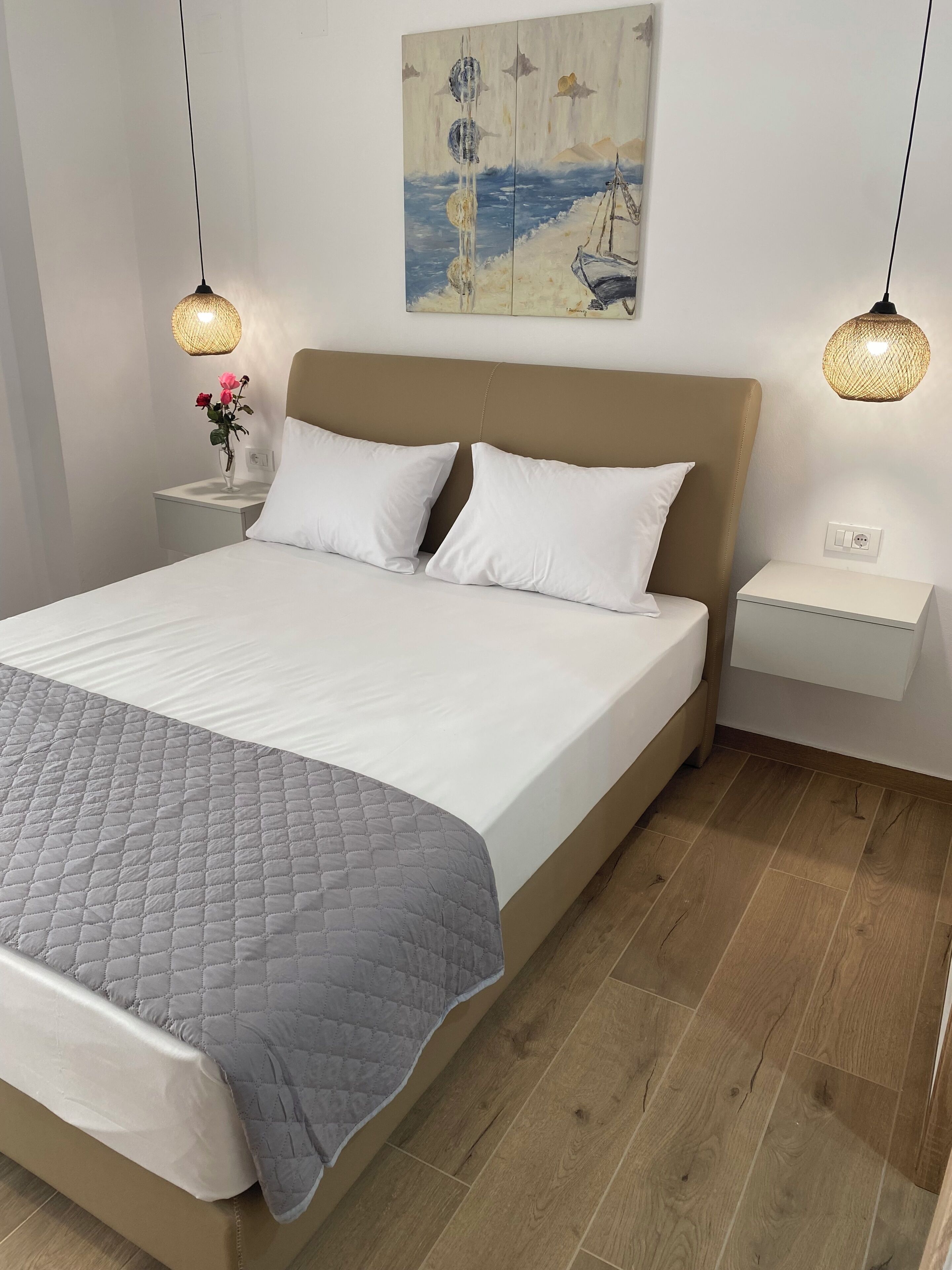 A neatly made bed with a quilt, two bedside tables with lamps, a painting on the wall, and a wooden floor.