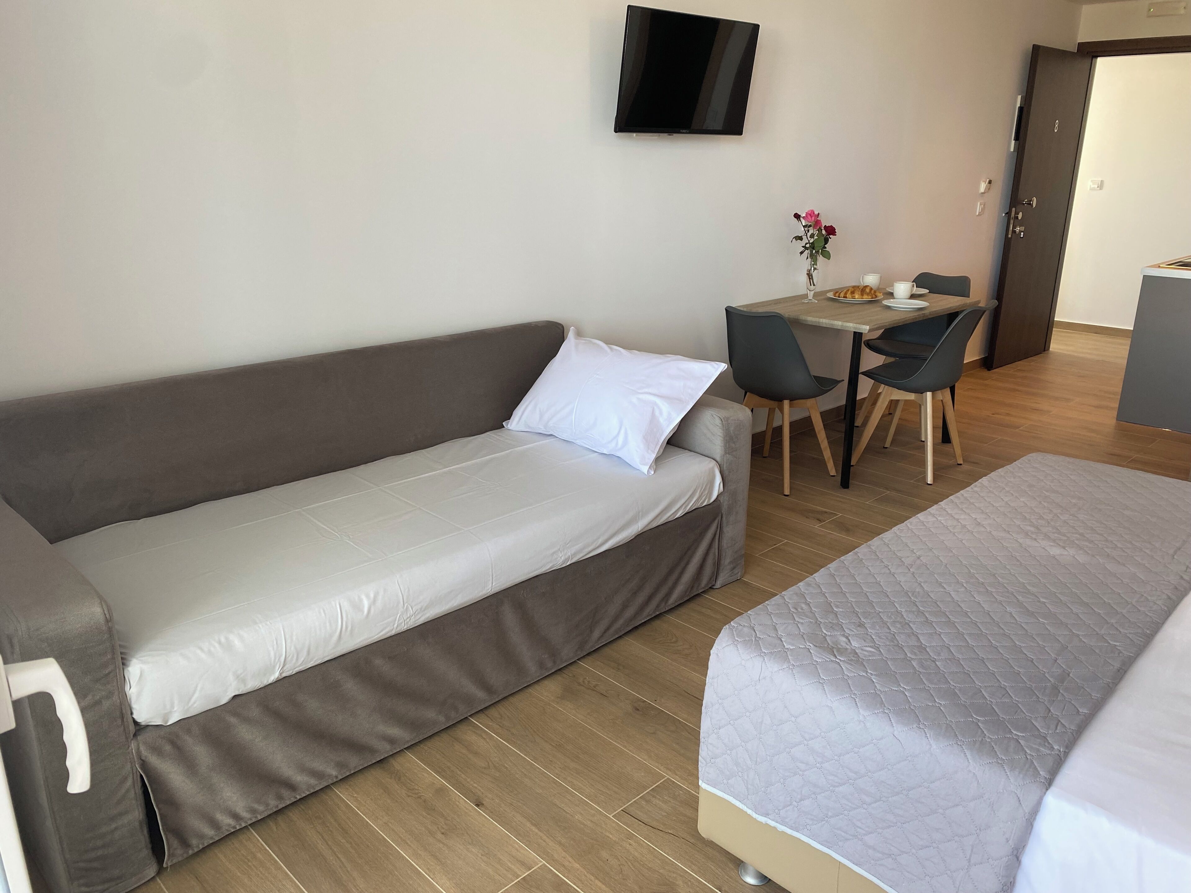 Een moderne hotelkamer met een bank, een kleine tafel met stoelen, een aan de muur bevestigde televisie en een deur.