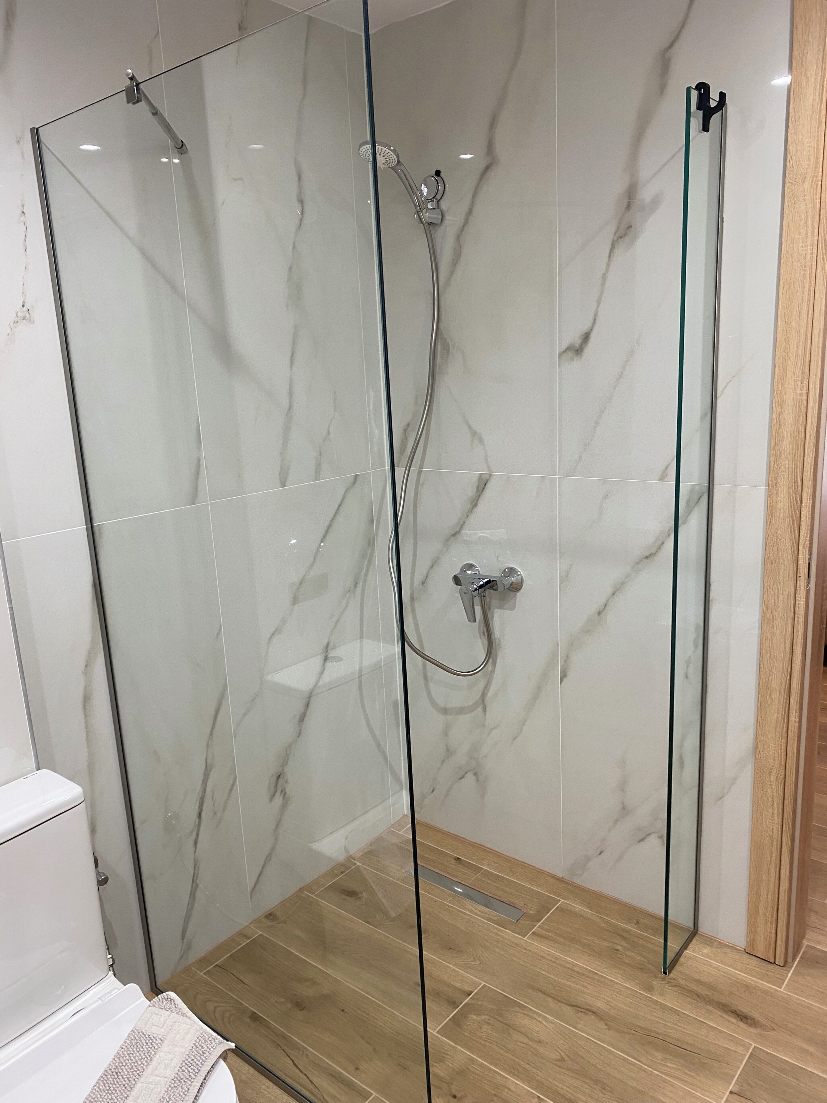 Eine Duschkabine mit Glastüren, eine Marmorwand, ein Holzboden und eine weiße Toilette.