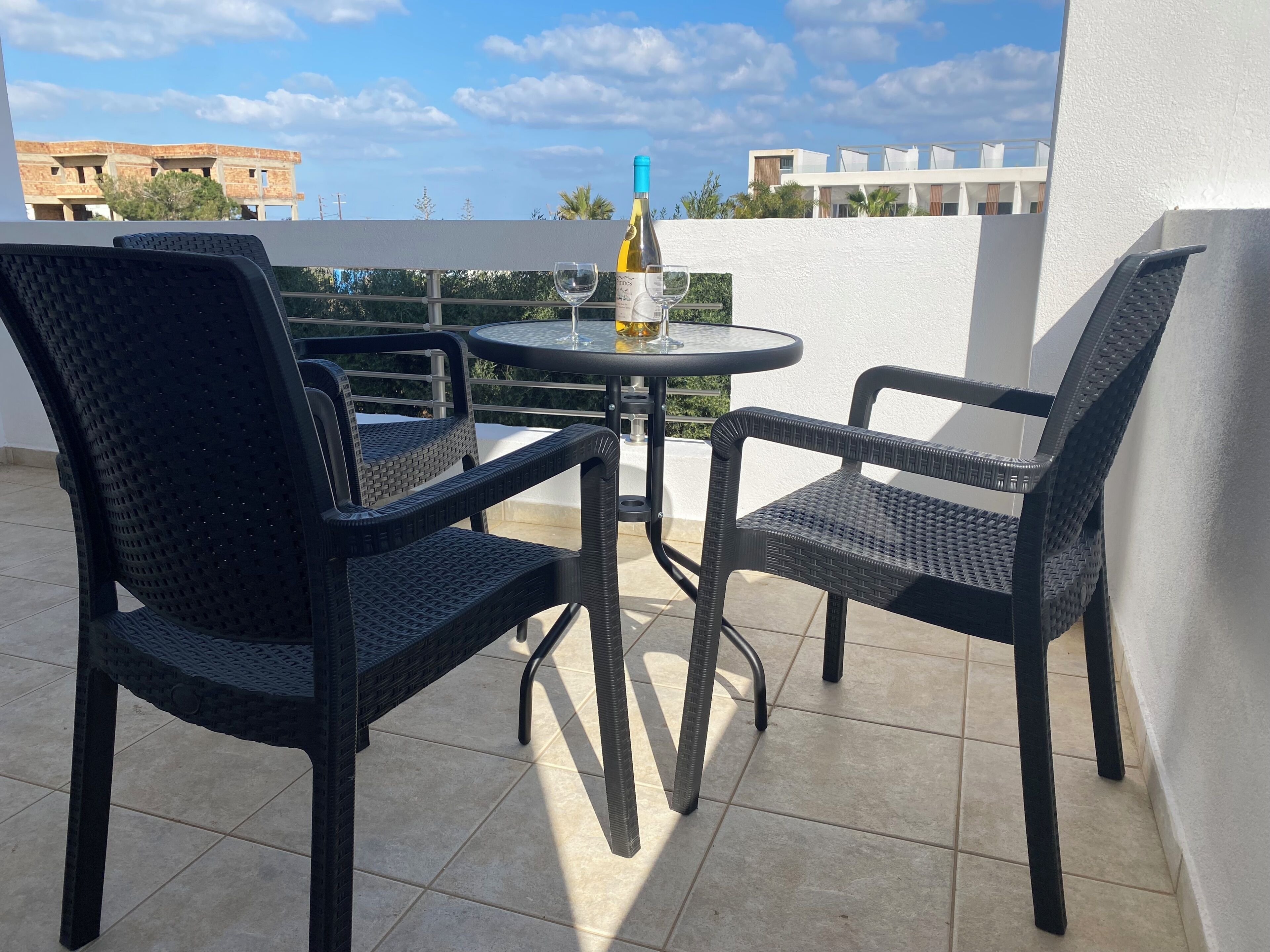 Terraço com duas cadeiras de vime e uma mesa, uma garrafa de vinho e céu claro.