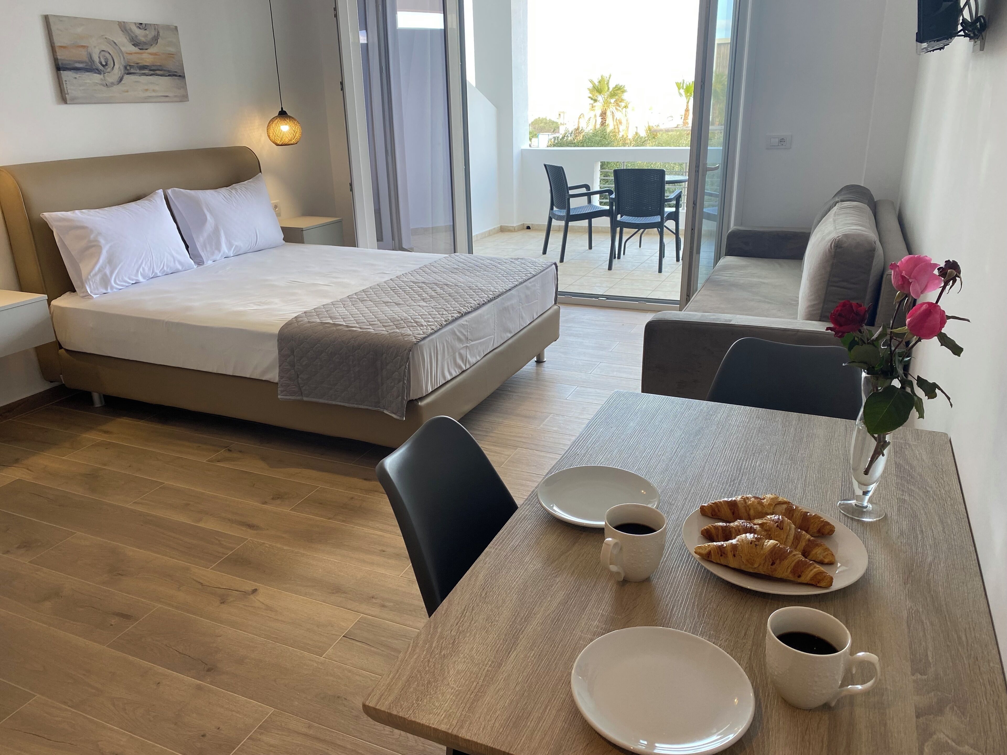 Une chambre d’hôtel avec un lit, une table à manger avec des croissants et du café, un vase rempli de fleurs et un balcon avec des chaises.