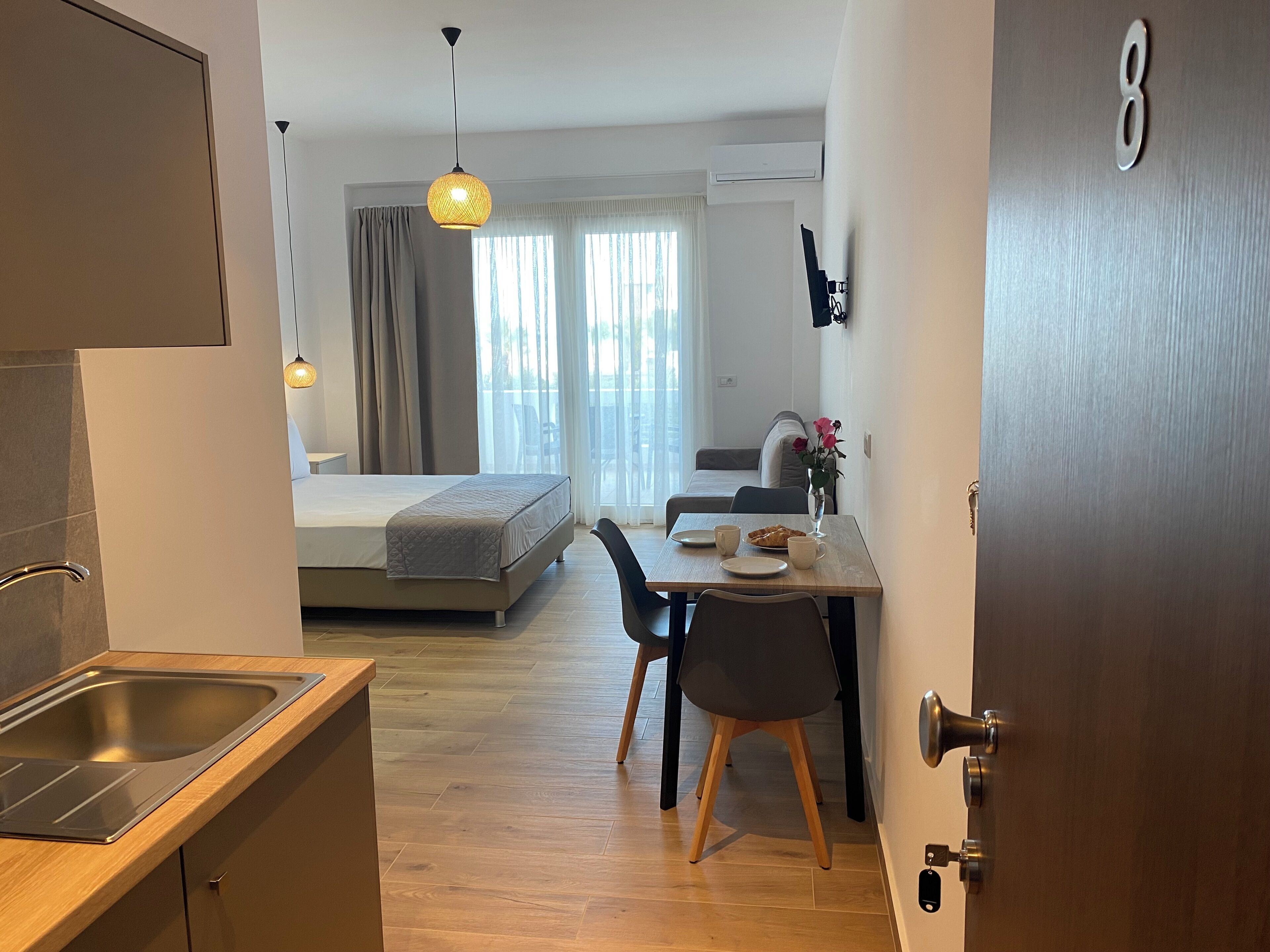 A modern hotel room with a kitchenette, a dining area with a table and chairs, a bed, and a view of the outdoors.