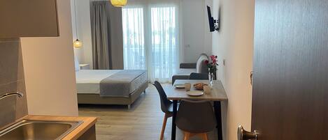 A modern hotel room with a kitchenette, a dining area with a table and chairs, a bed, and a view of the outdoors.