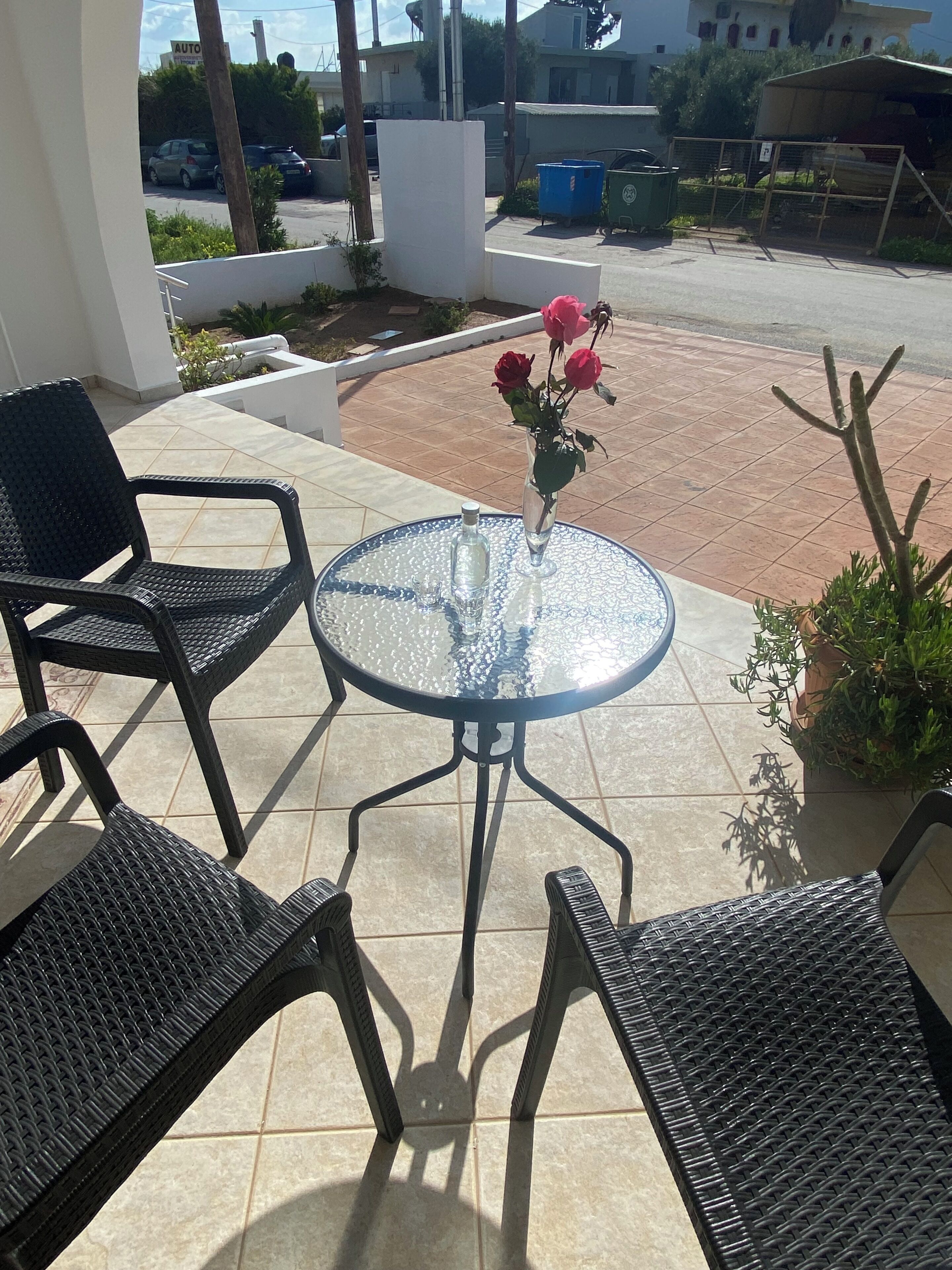 A patio with a round glass table, two black wicker chairs, and a vase with red roses.