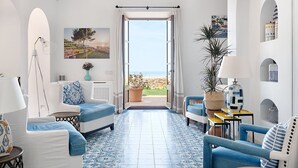 Un salón amplio con vista al mar, que incluye un sofá blanco, sillones azules y una mesa de centro.