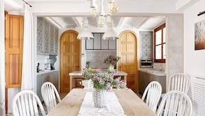 Un comedor con una mesa larga, sillas blancas, un florero con flores y una cocina al fondo.