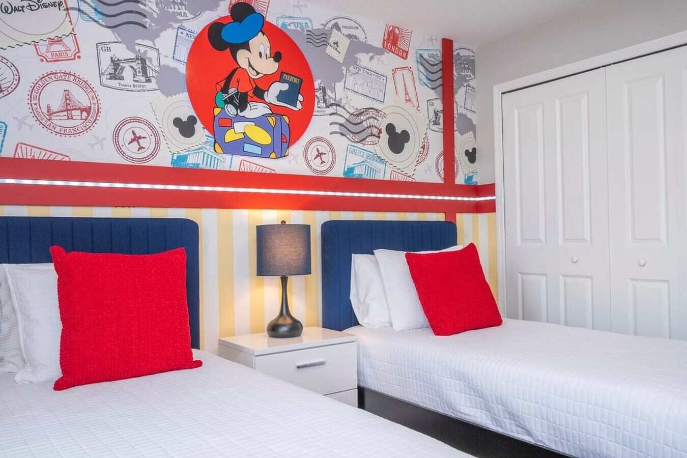 Une chambre d’hôtel avec deux lits, une table de chevet, une lampe et un mur décoré de timbres de voyage et d’une illustration de Mickey Mouse.