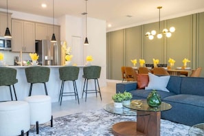 Cozinha moderna com armários brancos, geladeira de aço inoxidável e micro-ondas. Há uma área de jantar com mesa e cadeiras, e uma sala de estar com sofá azul e mesa de centro de vidro.