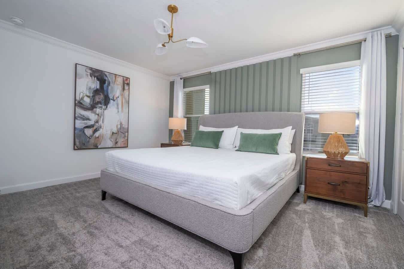 Een slaapkamer met een groot bed, twee nachtkastjes, een plafondlamp, een raam met jaloezieën en een schilderij aan de muur.