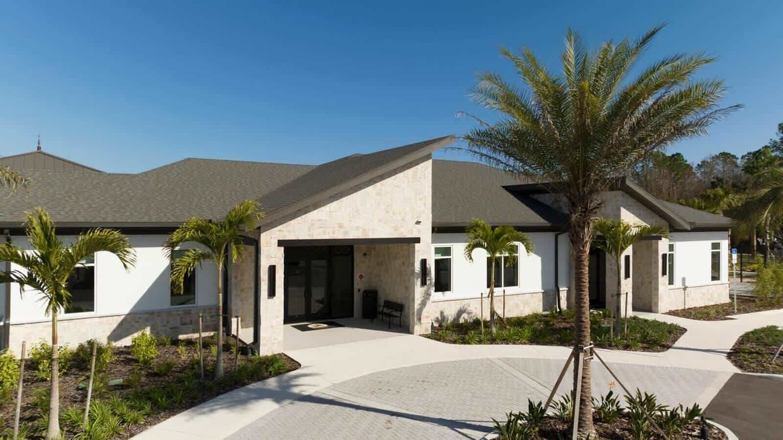 A modern two-story house with a stone facade, a covered patio, and a landscaped garden with palm trees.