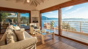 A spacious living room with a large window overlooking a body of water and mountains. There is a sofa, a coffee table, and a dining area visible.