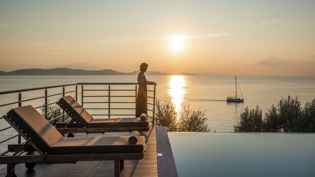 A sunset view from an infinity pool with lounge chairs and a person standing by the railing.