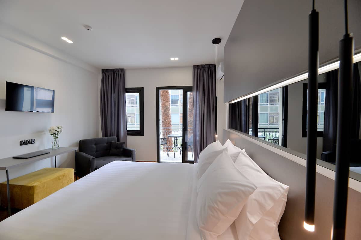 Habitación de hotel con una cama grande, un escritorio con televisor de pantalla plana, una silla y un balcón con mesa y sillas.
