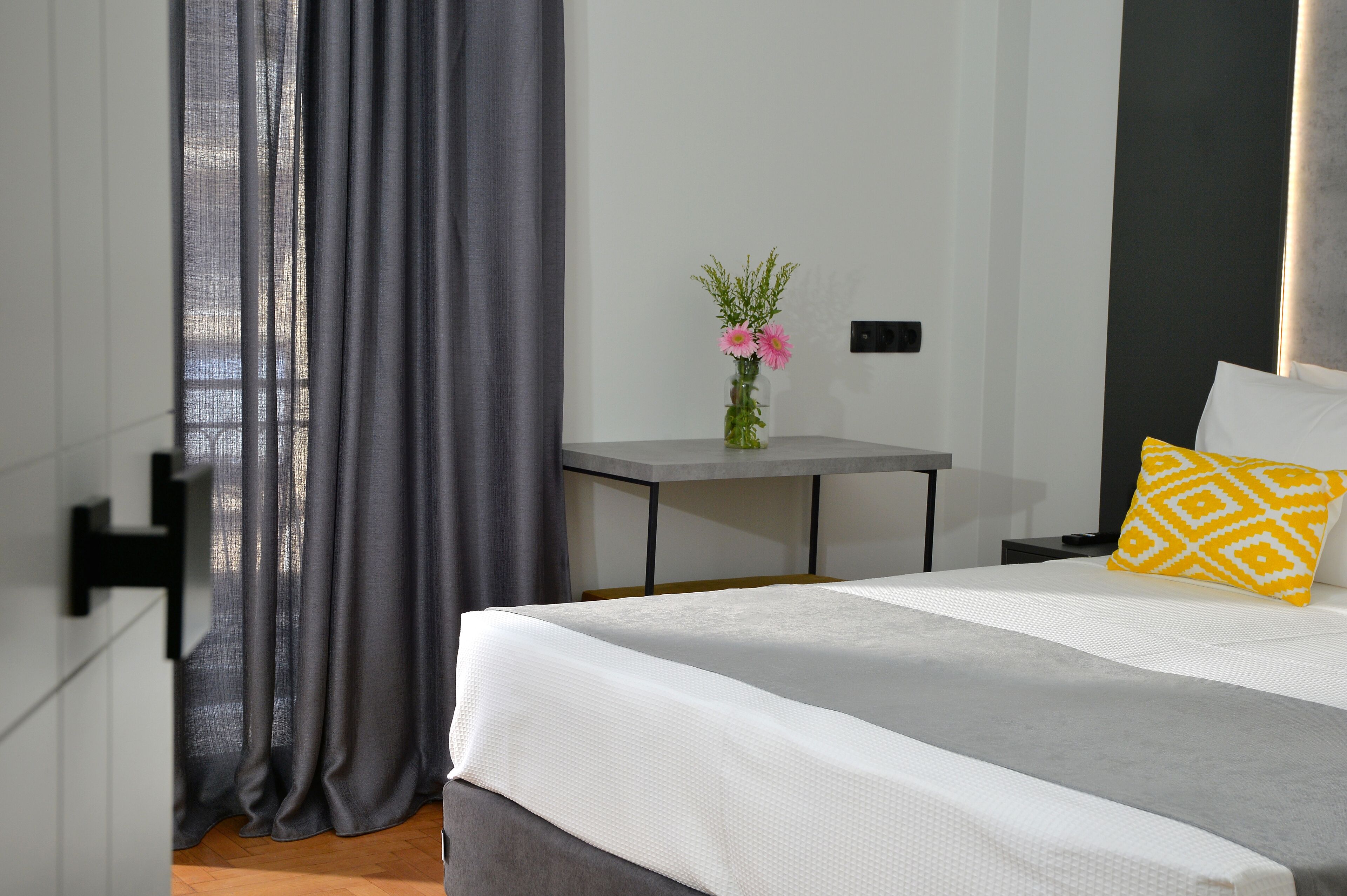A modern bedroom with a bed, a bedside table with a vase of flowers, and a view through a window with curtains.