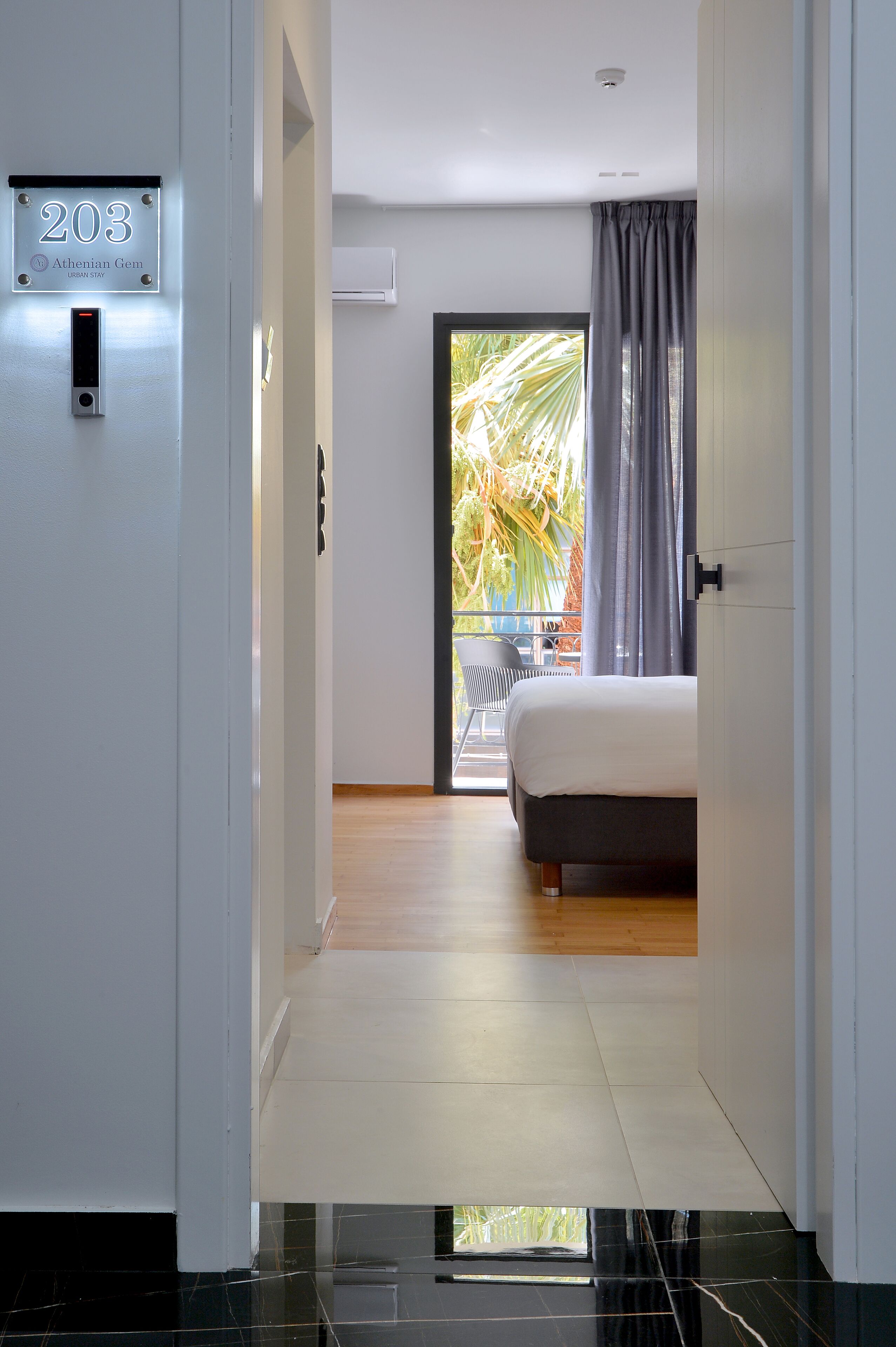 A modern hotel room with a bed, a balcony with a view of palm trees, and a door with a number sign.