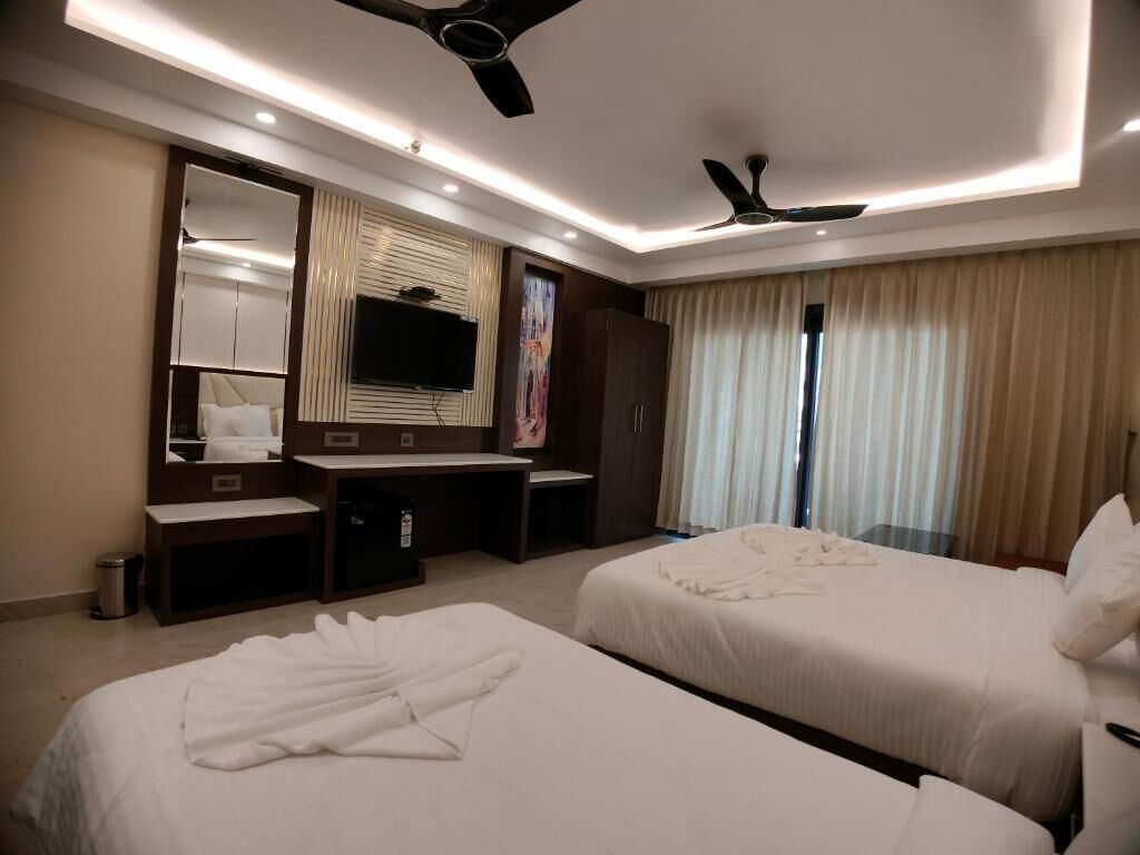 A hotel room with two beds, a desk, a mirror, a ceiling fan, and a large window with curtains.