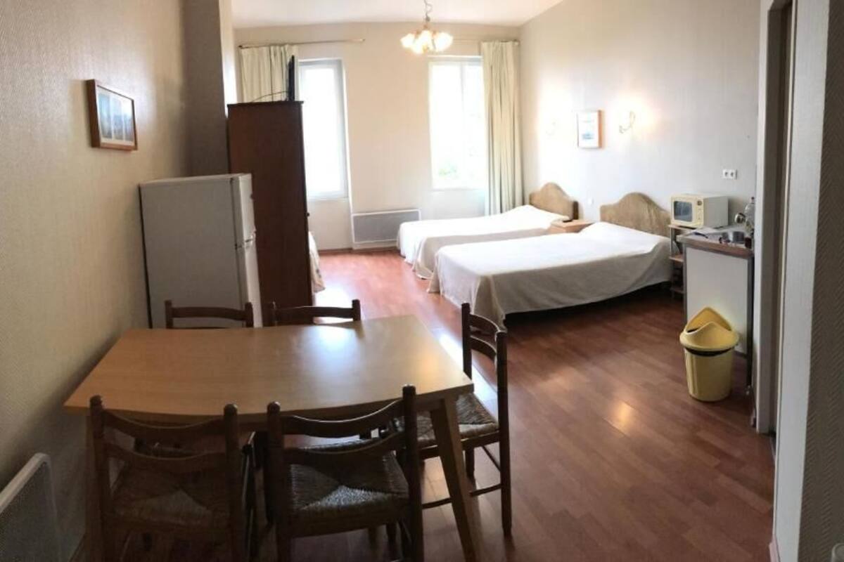 A room with a wooden dining table and chairs, two single beds, a small kitchenette, and a window with curtains.