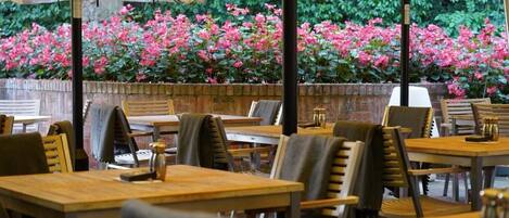 Un área de comedor al aire libre con mesas y sillas, rodeada por un muro de ladrillo y un jardín con flores rosadas.
