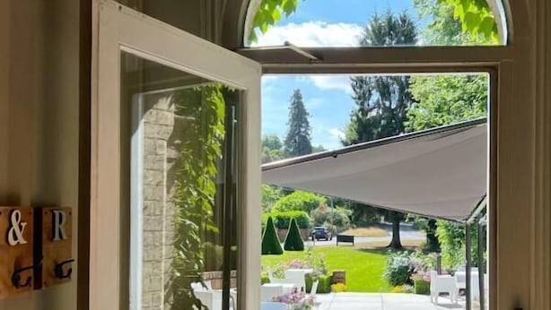 An open door leading to a covered outdoor patio with a white table and chairs, a garden view, and a decorative chandelier.