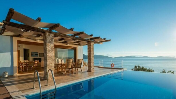 Una piscina infinita con vista al mar, un área de estar cubierta con sillas y un pilar de piedra.