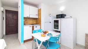 Een kleine keuken met witte kastjes, een magnetron en een koelkast. Er staat een eettafel met blauwe stoelen en een klein ontbijtarrangement.