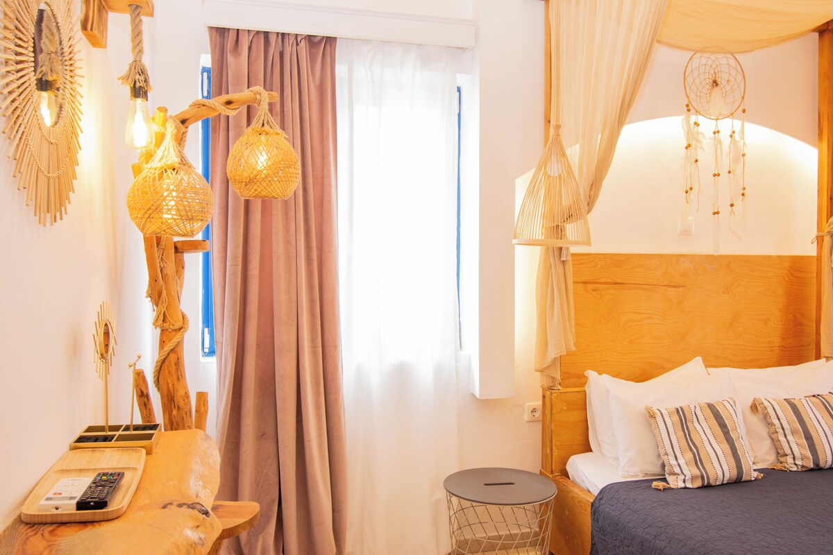 A room with a bed, a bedside table with a remote control, a round side table, a hanging dreamcatcher, and a window with curtains.