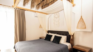 A neatly made bed with a grey bedspread, a wooden headboard, and two black pillows. There is a wooden nightstand with a drawer and a hanging dreamcatcher on the wall. - Ov Suites (Hersonissos)