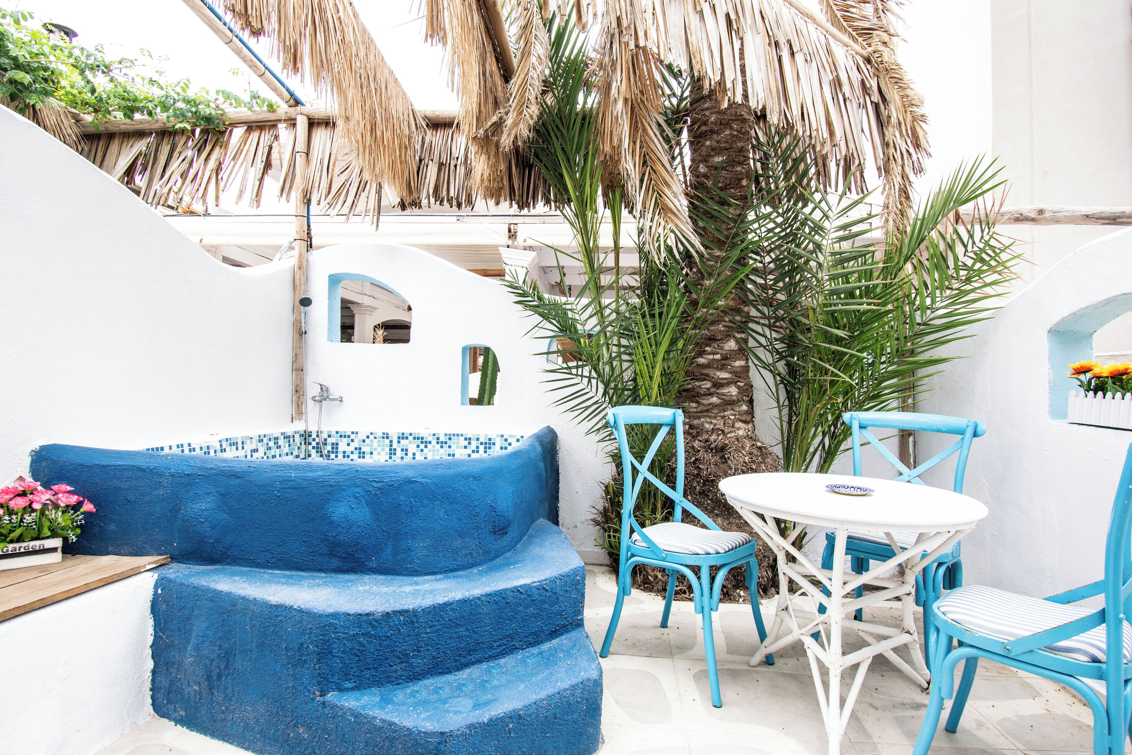A blue and white outdoor seating area with a thatched roof, palm trees, and a white wall with a door.