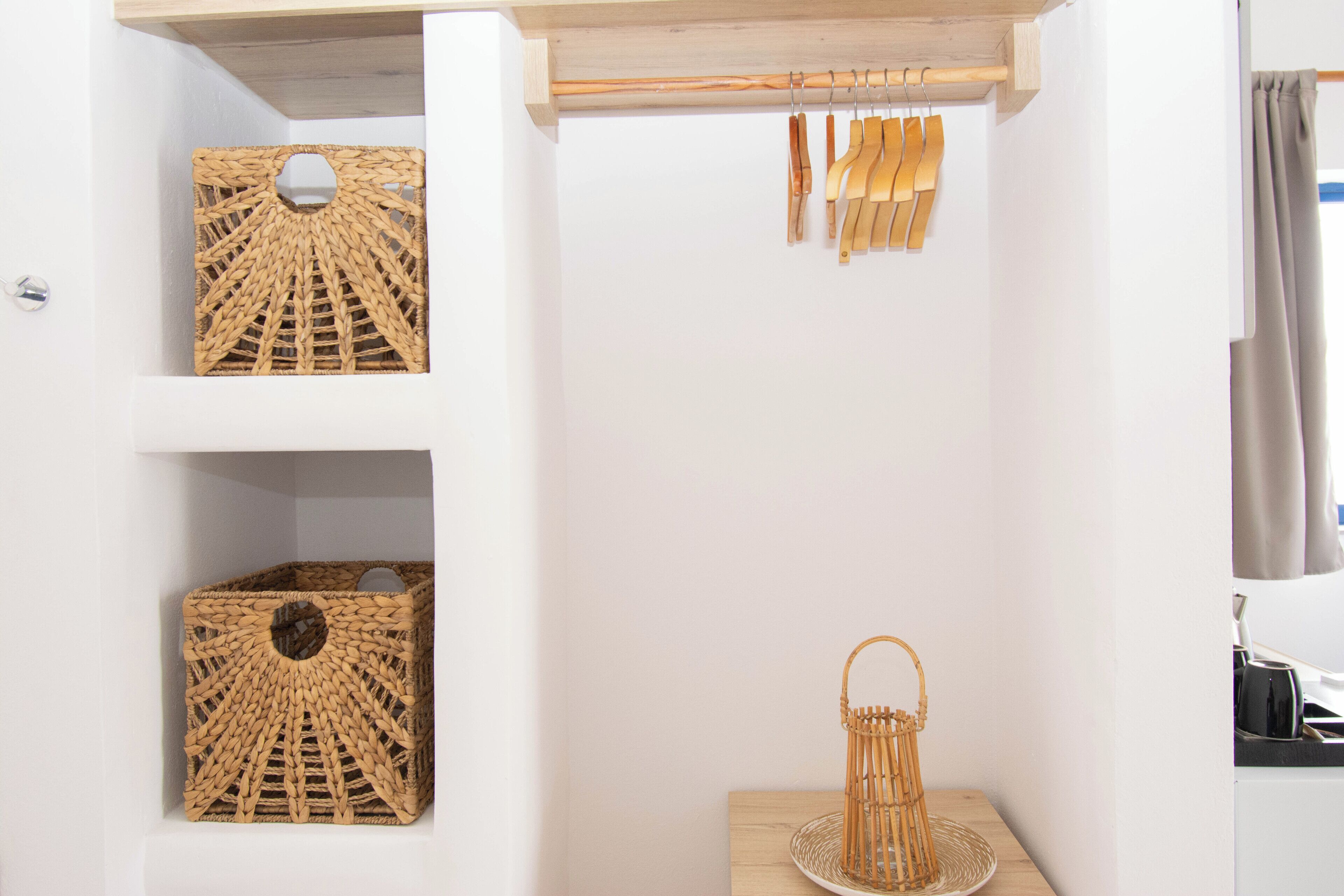 Ein Zimmer mit weißen Wänden, einem Holzregal mit gewebten Körben, einem Holz-Kleiderständer mit Kleidung und einem kleinen Tisch mit einem gewebten Korb.