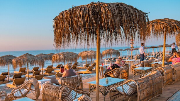 A beachside restaurant with thatched umbrellas, wicker furniture, and tables overlooking the ocean. - Ov Suites (Hersonissos)