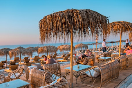 A beachside restaurant with thatched umbrellas, wicker furniture, and tables overlooking the ocean.