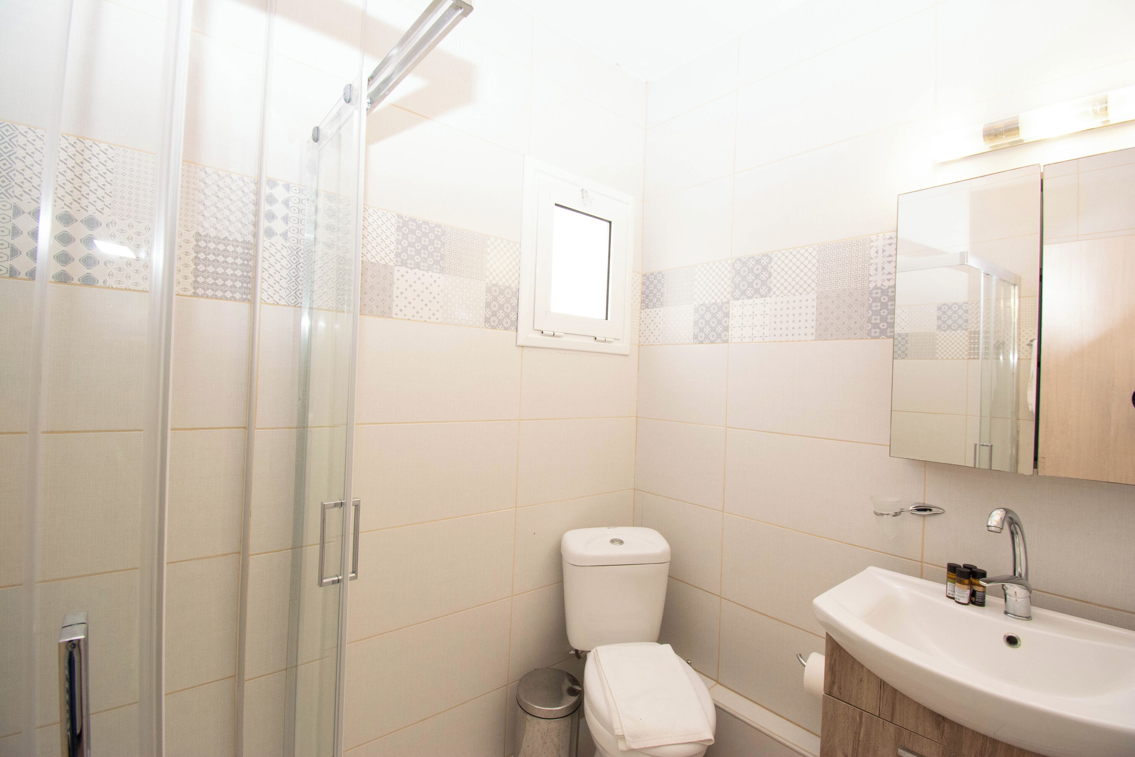 A bathroom with a glass shower enclosure, a toilet, a sink with a mirror above it, and patterned tiles on the walls.