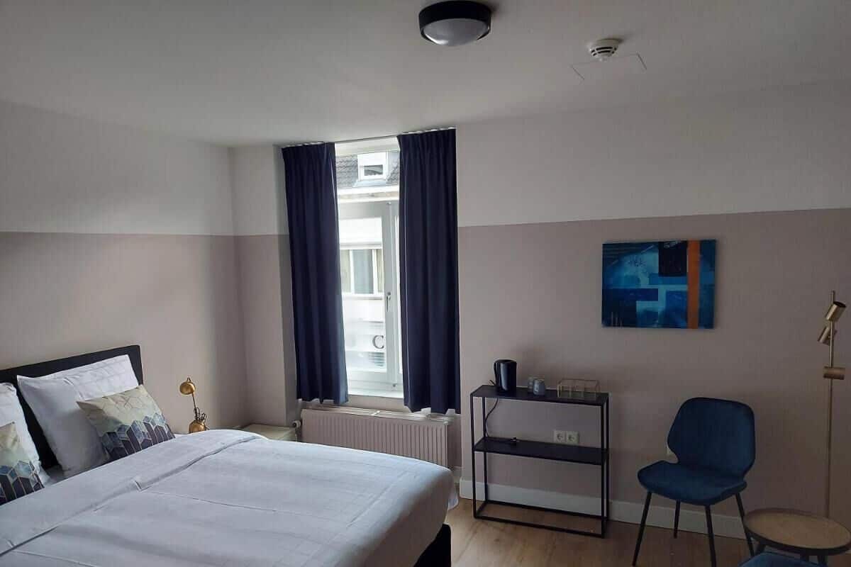 A hotel room with a bed, a desk, a chair, a radiator, and a window with curtains.