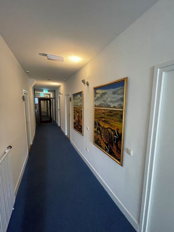 A hotel hallway with a blue carpet, white walls, and framed paintings on the right side. - Hotel Courage Valkenburg (Valkenburg aan de Geul)