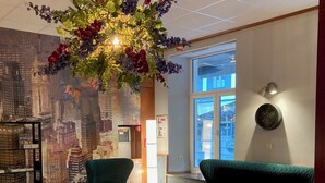 Un vestíbulo moderno con un mural de la ciudad, asientos tapizados en tonos verdes, un arreglo floral decorativo y un reloj redondo en la pared.