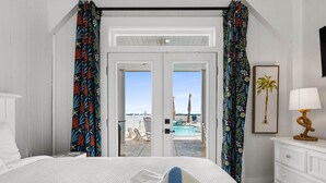 A hotel room with a bed, a white dresser, a lamp, a framed picture, and a view of a pool and palm trees.