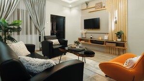 A modern living room with a black sofa, an orange armchair, a coffee table, and a TV mounted on the wall. - Kashco Apartment Wuse II (Abuja)