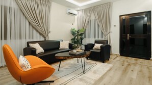 A modern living room with a black sofa, an orange chair, a coffee table, and a potted plant. - Kashco Apartment Wuse II (Abuja)