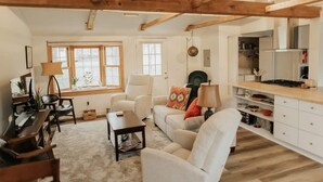 A cozy living room with a sofa, armchairs, a coffee table, and a kitchen area with cabinets and a stove. - Charming IU Cottage: Near Campus, Sleeps 8! (Bloomington)